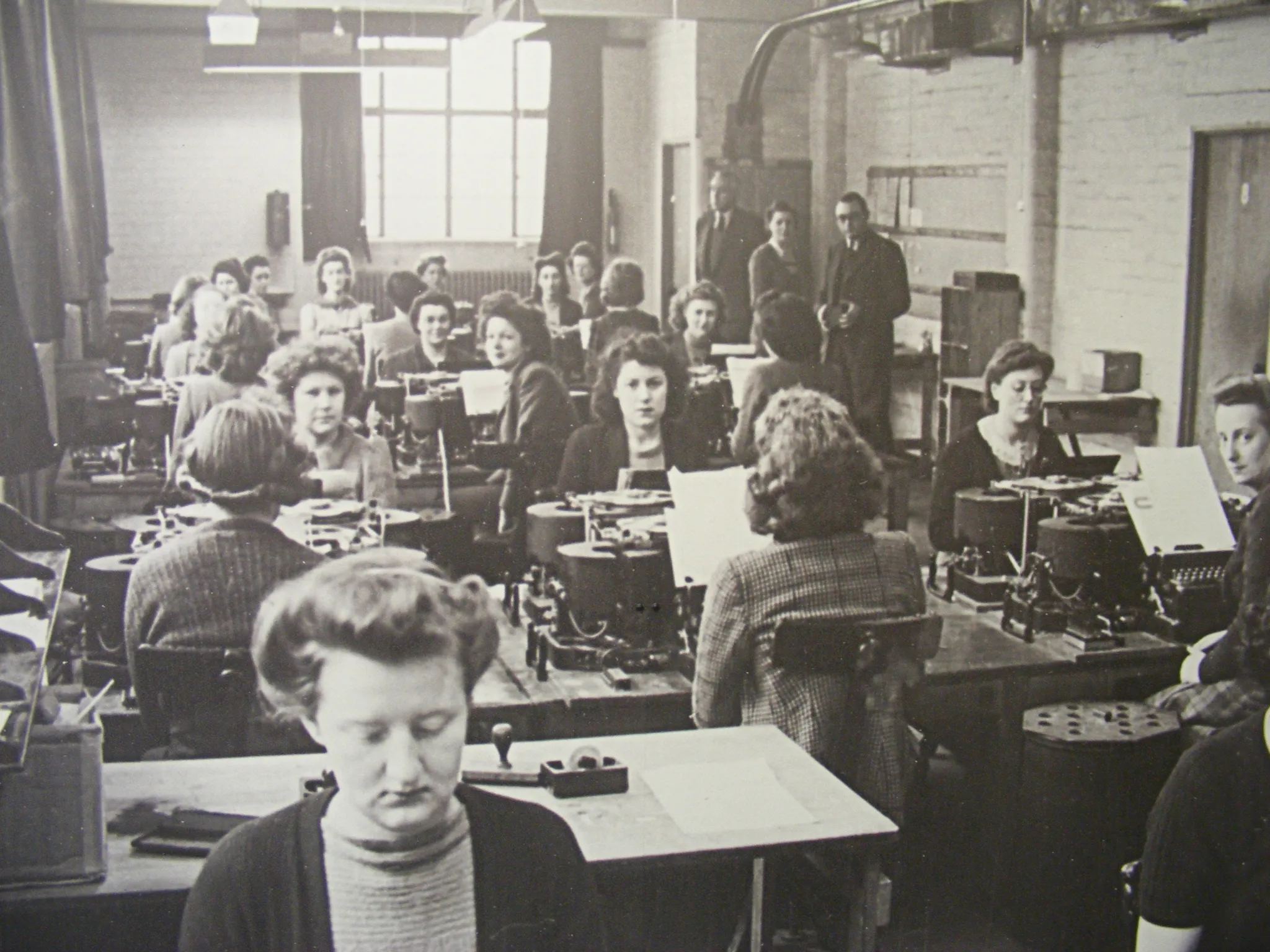 another black and white photo, this one shows many woman, over 10, sitting at desks arranged back to back, more details below